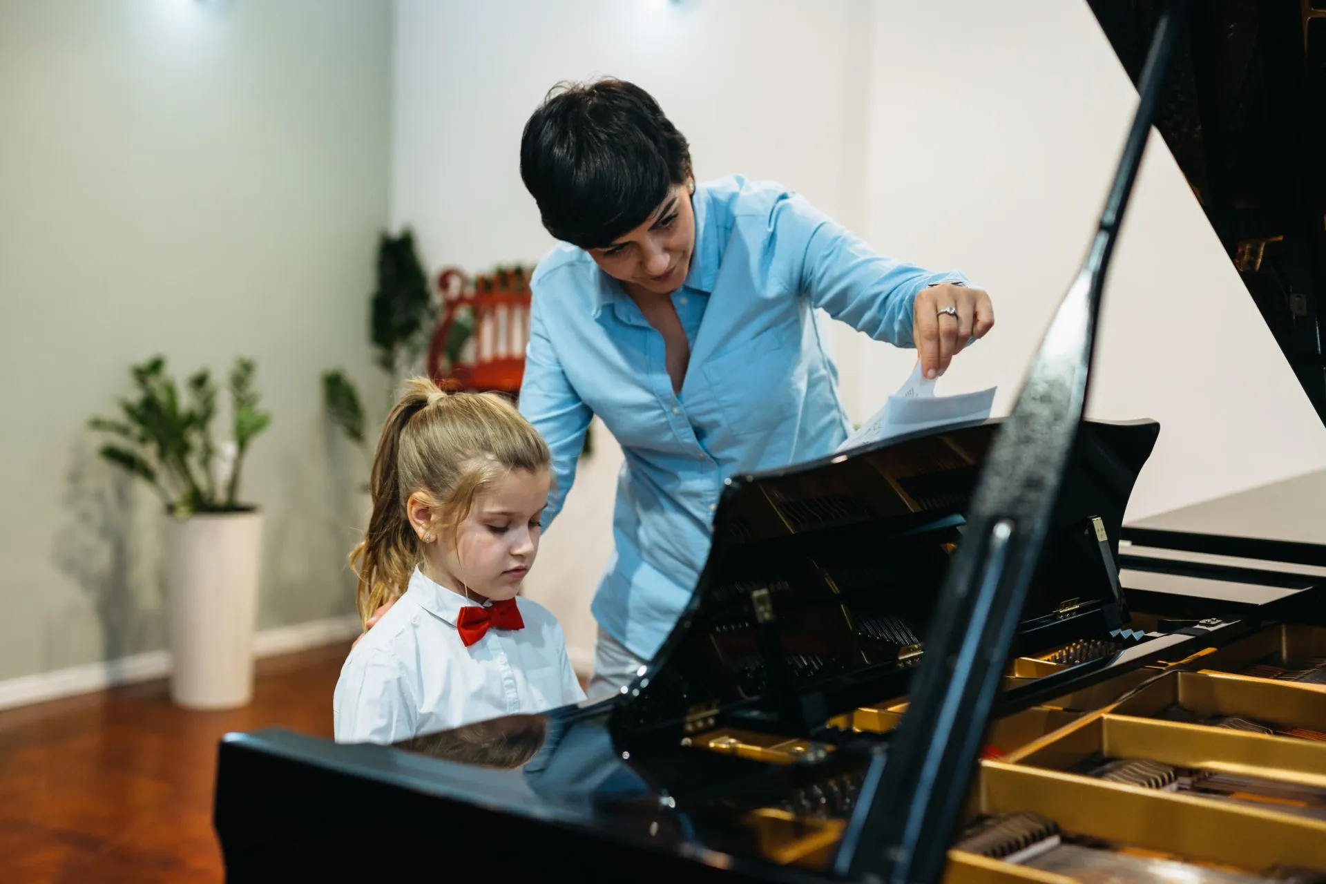 Piano teacher guiding a young girl in a red bow tie during a music lesson.