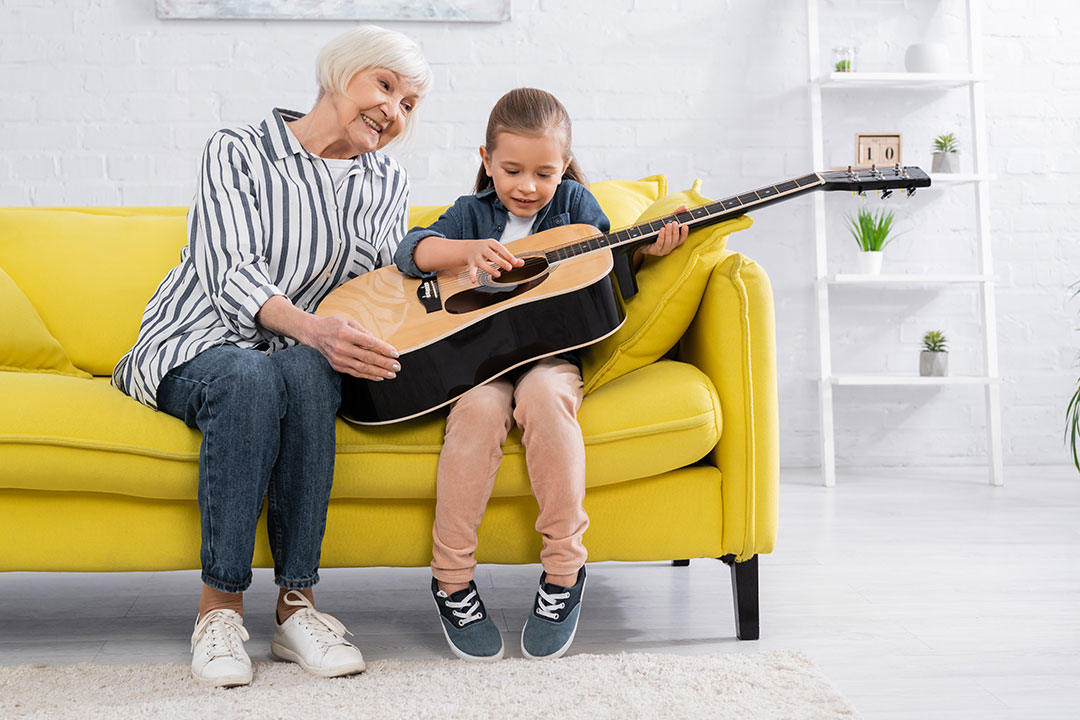 Grandmother and granddaughter playing guitar together on a yellow sofa, learning music at home | Learn The Art
